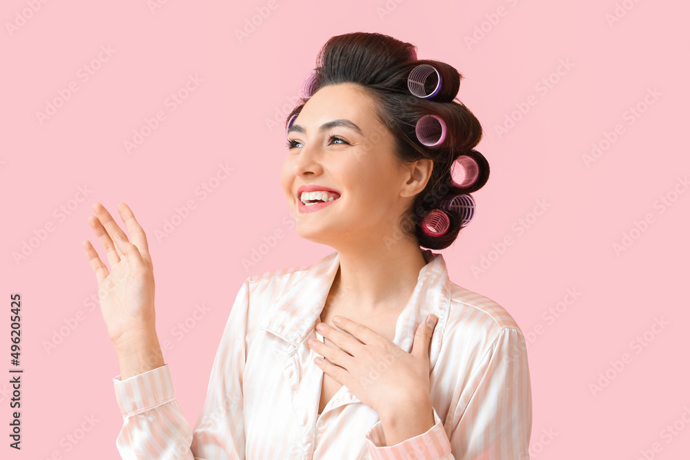 Beautiful young woman in pajamas and hair curlers on pink background