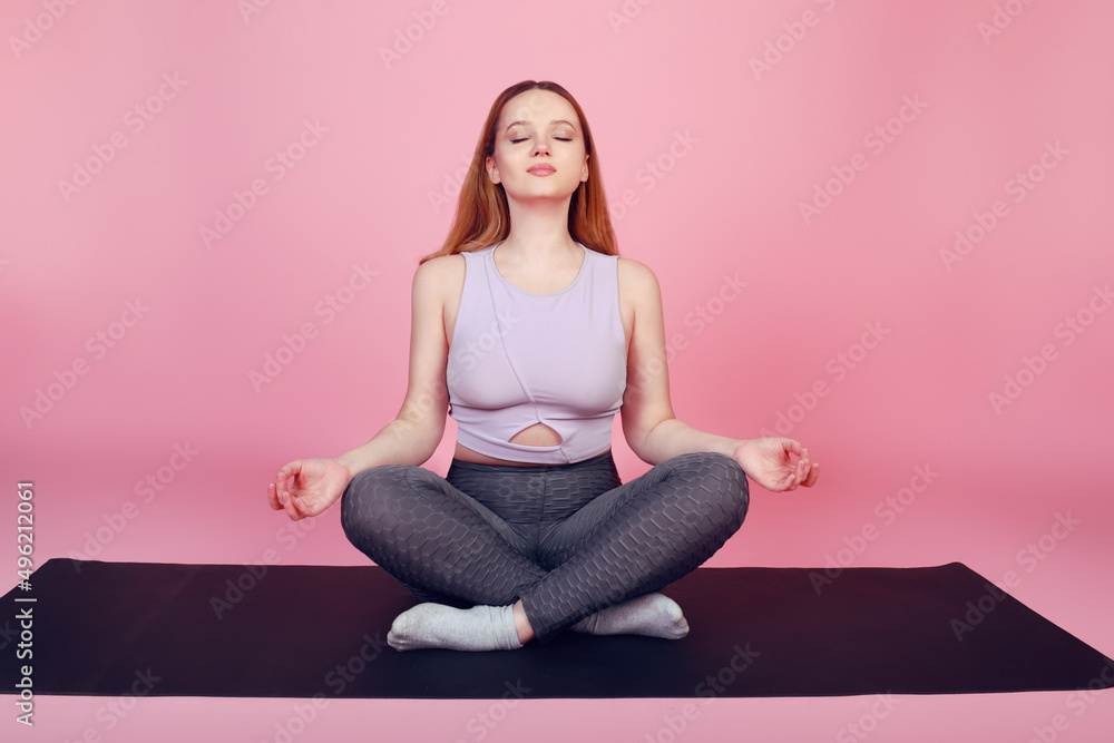 Beautiful body-positive girl on a pink background in a tracksuit goes in for sports, warm-up fitness sports, sits on a yoga mat