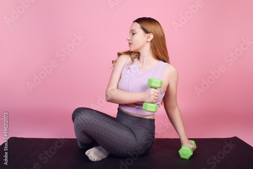 A beautiful body-positive girl on a pink background in a tracksuit goes in for sports, weights for training, exercises, fitness sports, yoga mat.