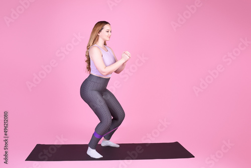 A beautiful body-positive girl on a pink background in a full-length sports suit goes in for sports, elastic bands for training, leg exercises, fitness