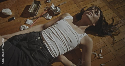 Wasted woman lying near drugs. From above female junkie lying on wooden floor near drugs and laughing in sunlight at home