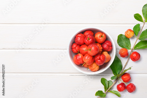 Flat lay of Acerola cherry in white bowl with branch on white wood.