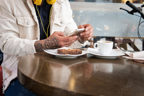 Close-up of the hands of an unrecognizable man who is typing a message on his cell phone at a coffee shop table next to a cup of coffee and a cake