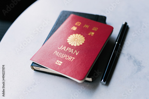 One single red Japanese passport laying on top of a black notebook next to a black pen that is on a white table