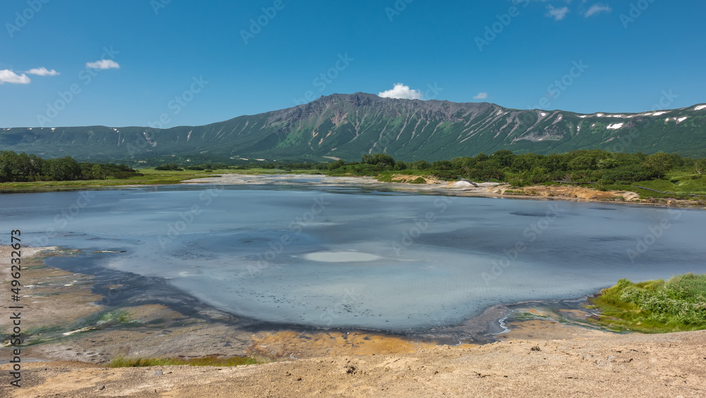 Blue lake in the caldera of an extinct volcano. Sulfur deposits are visible on the shores. Green vegetation in the distance. A picturesque mountain range against a clear sky. Kamchatka. Uzon