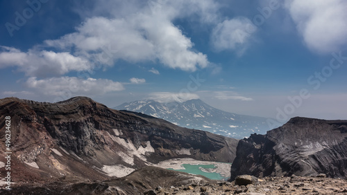 Turquoise acid lake on top of an active volcano. Melted snow at the water s edge. The layered structure of steep mountain slopes is visible. Clouds in the blue sky. Kamchatka. Gorely Volcano