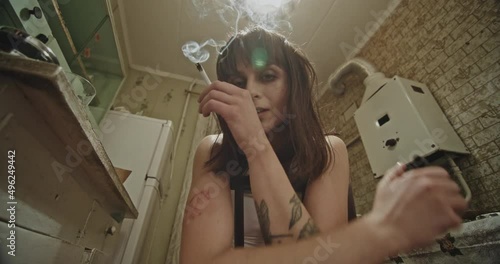 Female addict igniting joint with lighter. From below high woman igniting marijuana joint and and looking at camera while sitting on chair in grungy kitchen at home photo