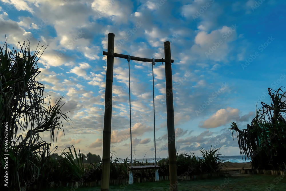 A  wooden swing with the view on Nyang Nyang Beach. The swing has very simple construction. There is green grass around the swing. Hidden getaway. Collecting happy moments.