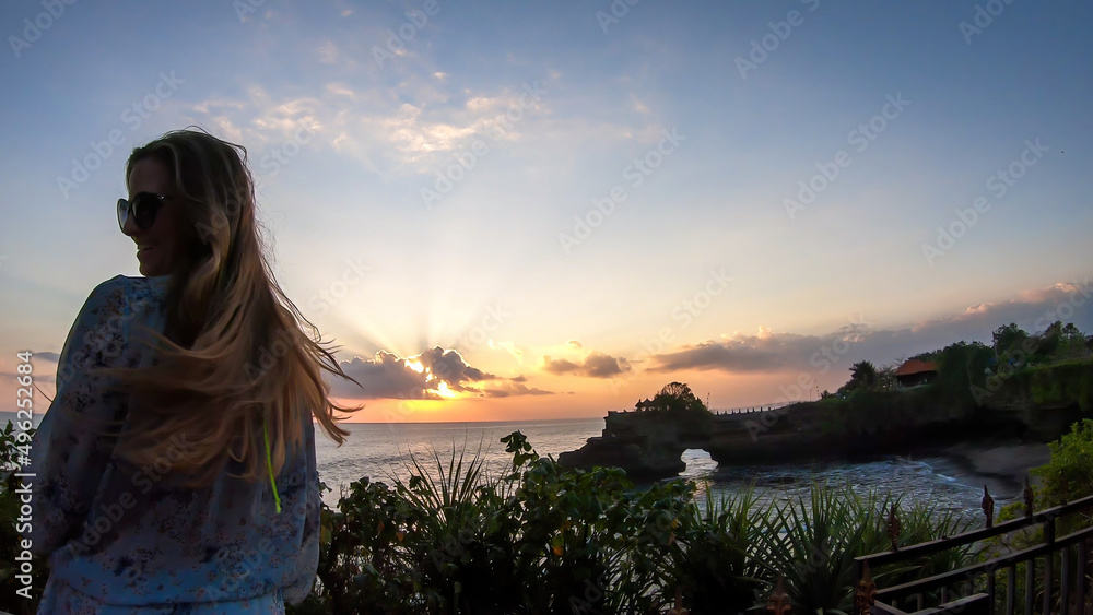 A close up on a woman is straw hat standing next to an edge of a cliffs, enjoying the early sunset hour in Tanah Lot Temple, Bali, Indonesia. Power of the nature. The woman is having a lot of fun