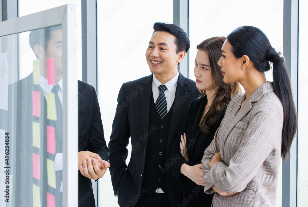 Asian happy male and female professional successful businessman and businesswoman colleagues partnership teamwork in formal business suit standing smiling look at post it note on glass board together Asian happy male and female professional successful businessman and businesswoman colleagues partnership teamwork in formal business suit standing smiling look at post it note on glass board together