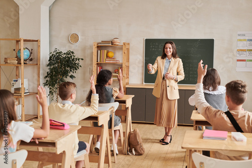 Wide angle view at young female teacher smiling while teaching class with group of children raising hands, copy space