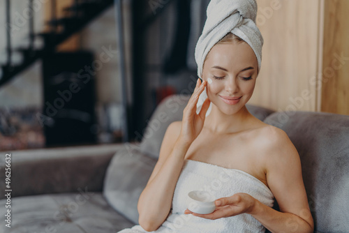 Beautiful female enjoys softness of skin afrer taking bath, applies moisturizing cream on cheek, wrapped in towel, sits on sofa alone