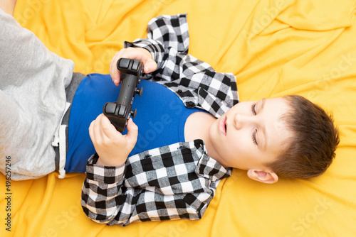 The child boy in checked shirt lying on the couch with black joystick in his hands playing the video game. Playing video games at home concept