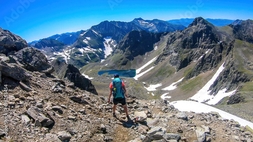 A young woman with a big backpack hikes down towards a clear, navy blue lake hiding between tall mountain peaks. Some of the slopes are covered with snow. In the back is another mountain range