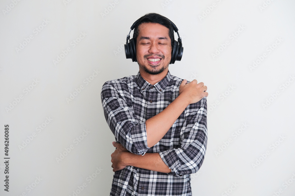 Fototapeta premium Adult Asian man showing relieved gesture when listening music using headset and hugging himself