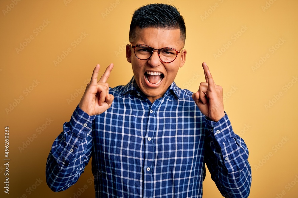 Young handsome latin man wearing casual shirt and glasses over yellow background shouting with crazy expression doing rock symbol with hands up. Music star. Heavy music concept. Young handsome latin man wearing casual shirt and glasses over yellow background shouting with crazy expression doing rock symbol with hands up. Music star. Heavy music concept.