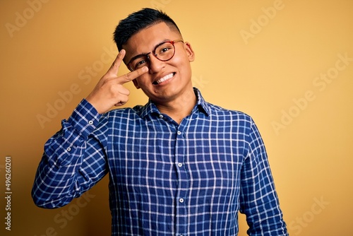 Young handsome latin man wearing casual shirt and glasses over yellow background Doing peace symbol with fingers over face, smiling cheerful showing victory