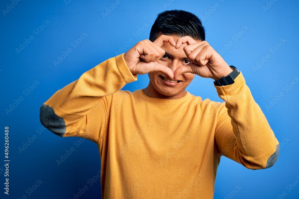 Naklejka premium Young handsome latin man wearing yellow casual sweater over isolated blue background Doing heart shape with hand and fingers smiling looking through sign