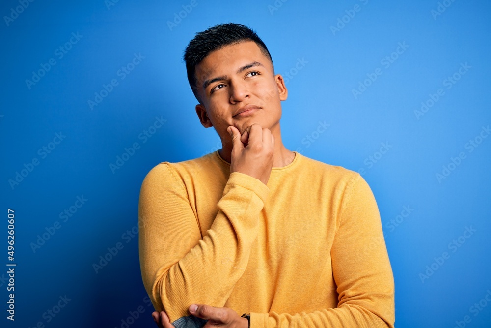 Young handsome latin man wearing yellow casual sweater over isolated blue background with hand on chin thinking about question, pensive expression. Smiling and thoughtful face. Doubt concept.