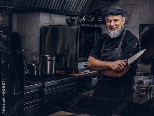 Joyful old man chef holding knife in modern kitchen