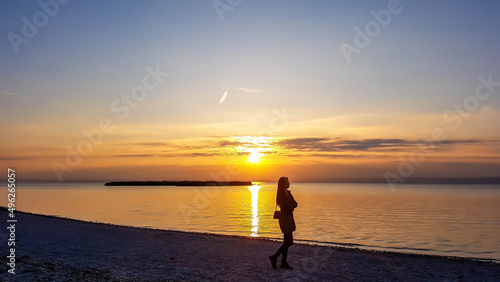 A young woman standing on the beach and watching sunset. The beach is pebbly and overgrown with grass. Sun reflects itself in the calm water of the lake. Sunrise above the lake. Freedom and happiness