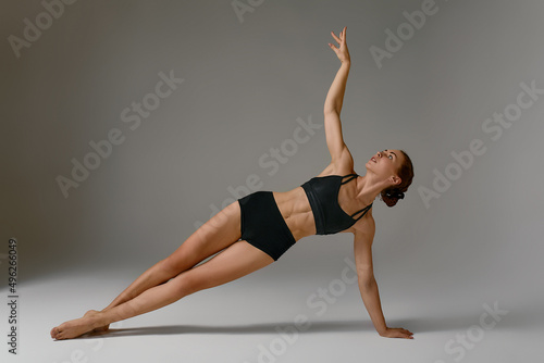 Beautiful flexible young gymnast woman in black leotard working out, stretching, doing backbend exercise, full length, studio, gray background
