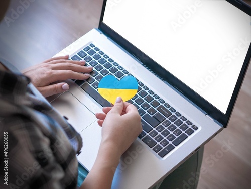 A brunette girl in a plaid shirt sits with a laptop holding a yellow and blue heart in front of the keyboard, the flag of Ukraine. Writes a letter to relatives or reads the news. Upper angle.