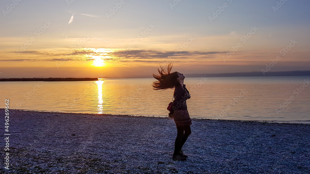 A girl standing at a pebbly beach by the lake and flipping her hair during the sunset. The hair are spreading all around the head of the girl. Sun is slowly reaching the lake. Freedom and happiness