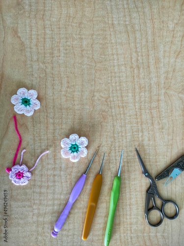 Crochet needles, scissors and crochet flowers on a light brown wooden background.