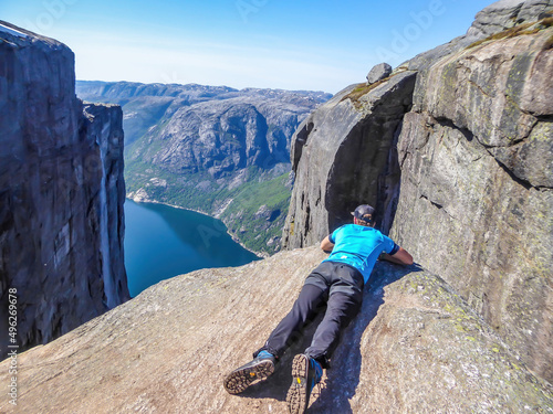 A young man lying at the edge of a steep mountain. He looks down into the valley. Overcoming the fear of heights. In front of her stunning Lysefjorden shimmering with many shades of blue and green