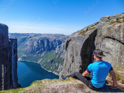 A young man sitting at the edge of a steep mountain. His legs are hanging down. Overcoming the fear of heights. In front of her stunning Lysefjorden shimmering with many shades of blue and green