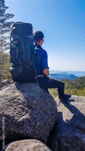 A young man with a huge hiking backpack sits at the rock and enjoys the view in front of him - fjords joining with the open sea. Camping in tall mountains. Adventure seeking. Clear and bright day.