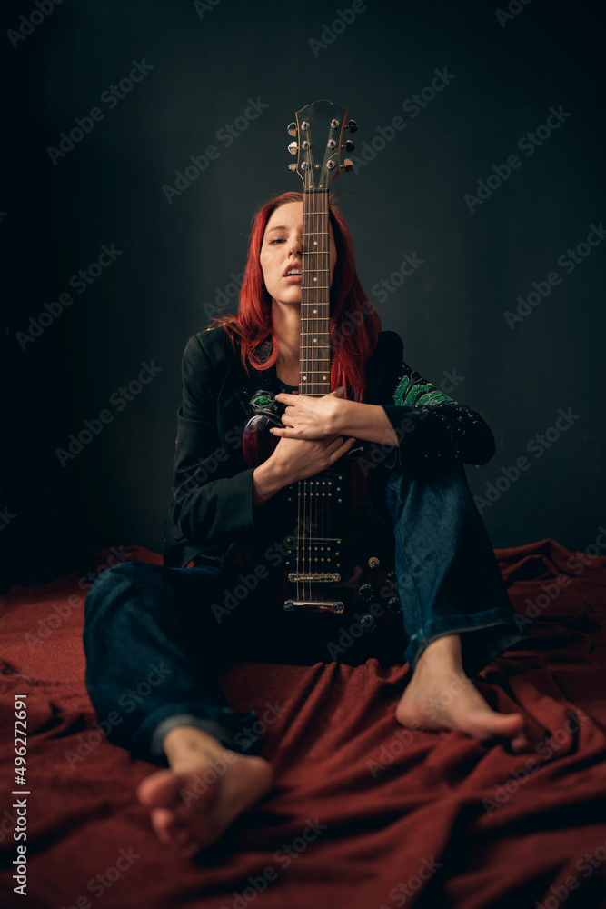 Sexy and brutal red-haired girl rocker, punk or metalhead, with an electric guitar in a black jacket and jeans sits on the floor in a defiant pose and hugs a guitar