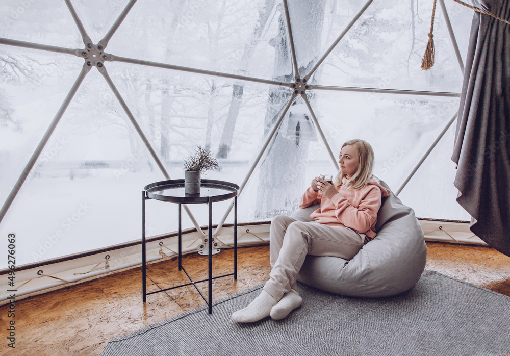 A woman is sitting in a geo dome glamping tent in a bag chair and looking out at the winter snow-covered nature and drinks coffee.