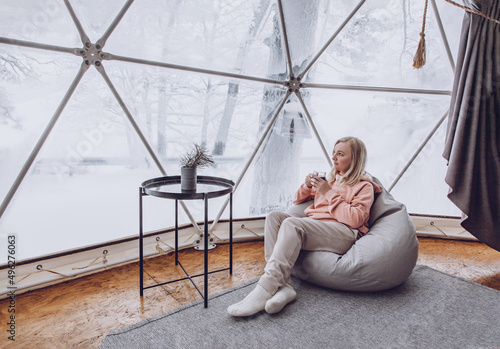 Obraz na płótnie A woman is sitting in a geo dome glamping tent in a bag chair and looking out at the winter snow-covered nature and drinks coffee. w salonie