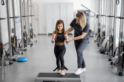 Rehabilitation specialist helping little girl to do exercises at gym. Concept of physical therapy for back health and correct posture for kids