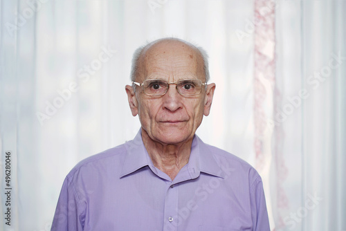 Portrait of hoary elderly man wearing eyeglasses standing at home against white transparent curtain