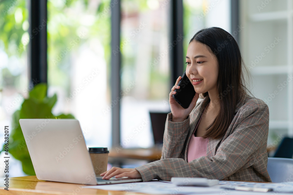 Portrait of young successful beautiful Asian businesswoman sitting at a desk at home office work on laptop talk on the cellphone. Smiling women use computers to have smartphone calls with clients. Portrait of young successful beautiful Asian businesswoman sitting at a desk at home office work on laptop talk on the cellphone. Smiling women use computers to have smartphone calls with clients.