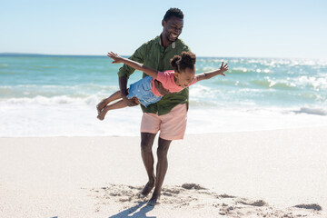 Full length of happy african american man carrying daughter pretending to fly at beach on sunny day