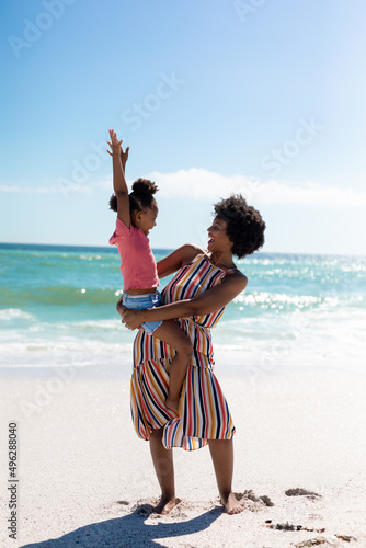 Cheerful african american mother carrying daughter while enjoying sunny day at beach against sky