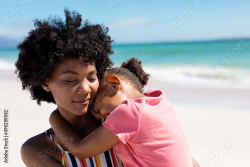 African american woman with eyes closed carrying daughter at beach on sunny day