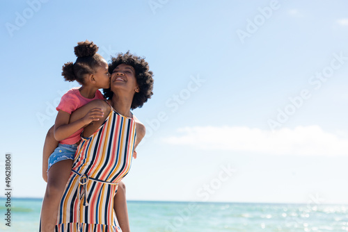 African american girl kissing mother while enjoying piggyback ride on her at beach against blue sky © wavebreak3