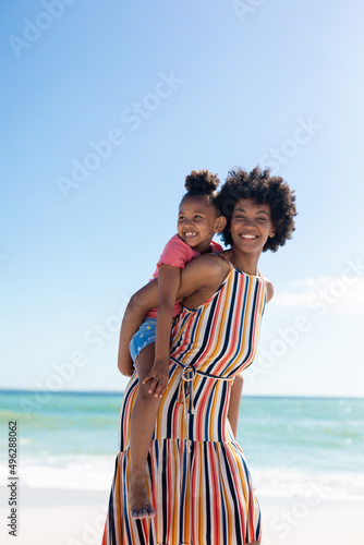 Portrait of happy african american mother giving piggyback ride to daughter at beach against sky
