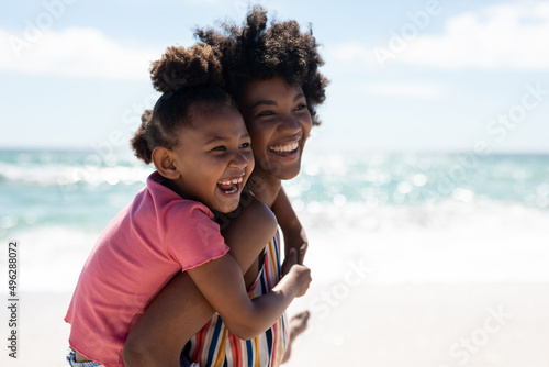 Happy african american mother giving piggyback ride to daughter at beach on sunny day