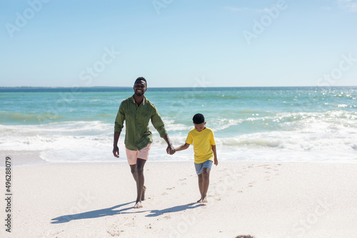 Full length of happy african american man holding hand of son while walking at beach against sky