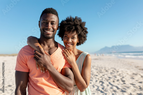 Portrait of smiling african american woman embracing boyfriend from behind at beach on sunny day