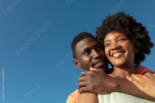 Low angle view of happy african american man hugging girlfriend from behind against clear blue sky