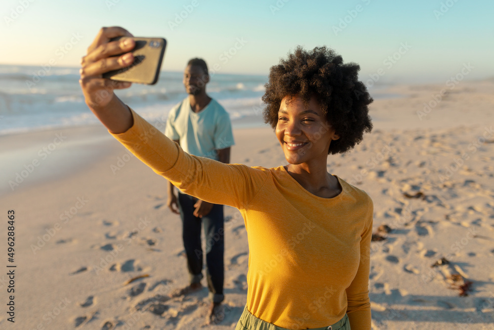 Happy african american woman taking selfie with man standing in background at beach