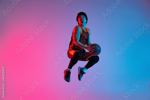 Young energetic man playing basketball isolated on gradient pink blue studio background in neon light. Youth, hobby, motion, activity, sport concepts.