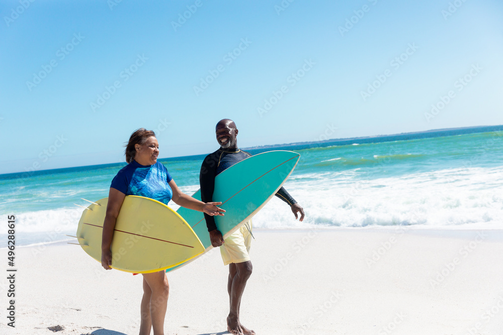 Fototapeta premium African american senior couple carrying surfboards walking at beach against blue sky with copy space